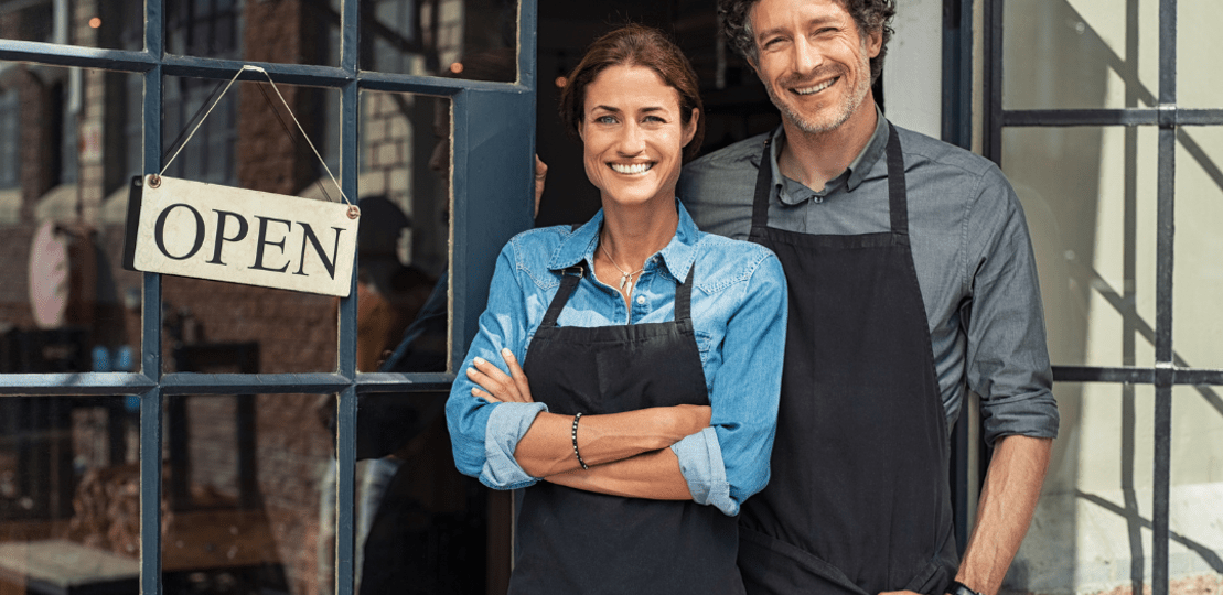 Woman and Man in front of small business with OPen sign hanging from door