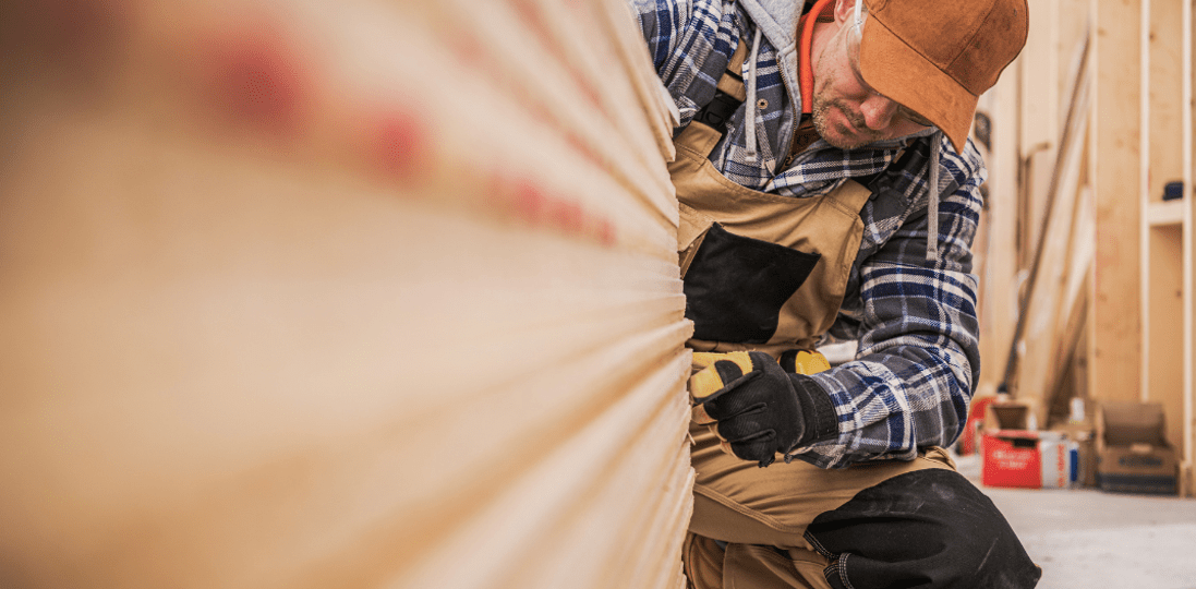 man in flannel shirt and carpenter pants with brown hat sanding a piece of wood